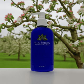 Vital Therapy Apple Delight Cleanser bottle in front of a blooming tree, offers soy-free, non-GMO skincare benefits.
