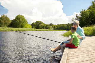 Older man and young boy fishing on a sunny day, sitting on a wooden dock by a serene lake surrounded by lush greenery.